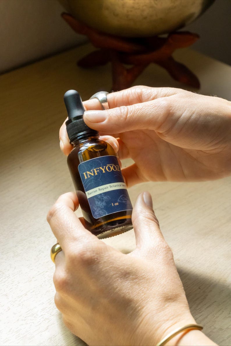 May include: A brown glass bottle with a black dropper is held between two hands. The bottle has a blue label with the text "INFYOOZ" and "Barrier Repair Botanical Serum". The label also indicates the bottle contains 1 oz of product.
