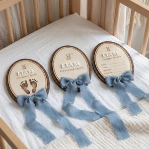 May include: Three wooden oval plaques with baby footprints, names, and blue ribbons. The plaques are in a wooden crib. One plaque says "Liam Alexander Smith". The other says "Liam" and the third says "Thomas Brown".