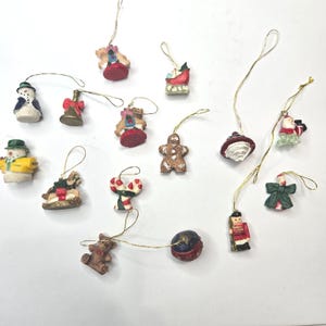 May include: A collection of fourteen miniature Christmas ornaments featuring various figures including snowmen, a gingerbread man, a teddy bear, a toy soldier, a sleigh, a bell, a candy cane, and a bow, each with a gold hanging string attached.