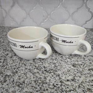 May include: Two off-white ceramic mugs with rounded bodies and side handles. Each mug features a black horizontal line with the text "CAPPUCCINO" and "Mocha" printed in a serif font. The mugs have a smooth, glossy finish and a wide opening.