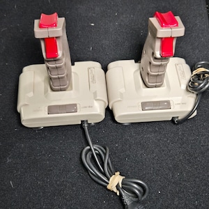 Puede incluir: Dos joysticks de juego de color beige con botones de disparo rojos y botones superiores rojos. Cada controlador cuenta con un agarre vertical, una base rectangular y un interruptor deslizante en la parte frontal. Cables negros unidos a cada unidad.