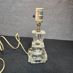 May include: A clear glass lamp base featuring a tiered, square-shaped design with a metallic socket at the top. The lamp includes a light-colored electrical cord extending from the side, designed for a standard light bulb and shade attachment.