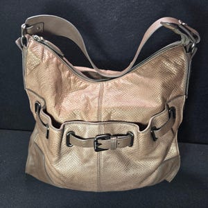 May include: A metallic bronze hobo bag with a textured snakeskin-like pattern. The bag features a single shoulder strap, a top zipper closure, and a front exterior pocket secured by a horizontal strap with a dark metal buckle and side hardware.