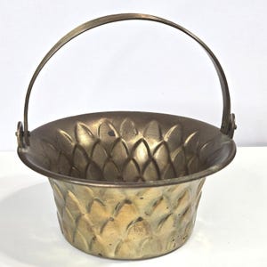 May include: A round brass bucket featuring a textured, embossed scale pattern across its exterior and interior surface. The vessel includes a thin, arched metal handle attached to two circular side mounts, showcasing a metallic gold finish.