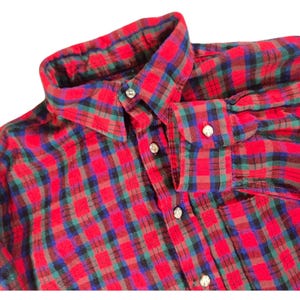 May include: A red, green, blue, and black plaid button-down shirt. The shirt has a classic collar and buttoned cuffs. The fabric appears to be a soft flannel. The shirt is a close-up shot on a white background.