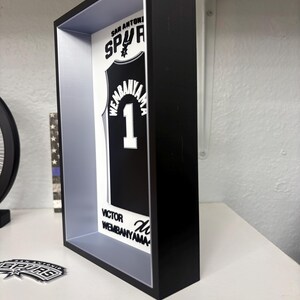 K&ouml;nnte beinhalten: Ein schwarzer Rahmen zeigt ein schwarz-wei&szlig;es San Antonio Spurs-Trikot mit dem Namen "Wembanyama" und der Nummer "1". Das Trikot wird in einer schwarzen Schattenbox mit wei&szlig;em und grauem Innenraum pr&auml;sentiert. Der Text "San Antonio Spurs" ist oben zu sehen.