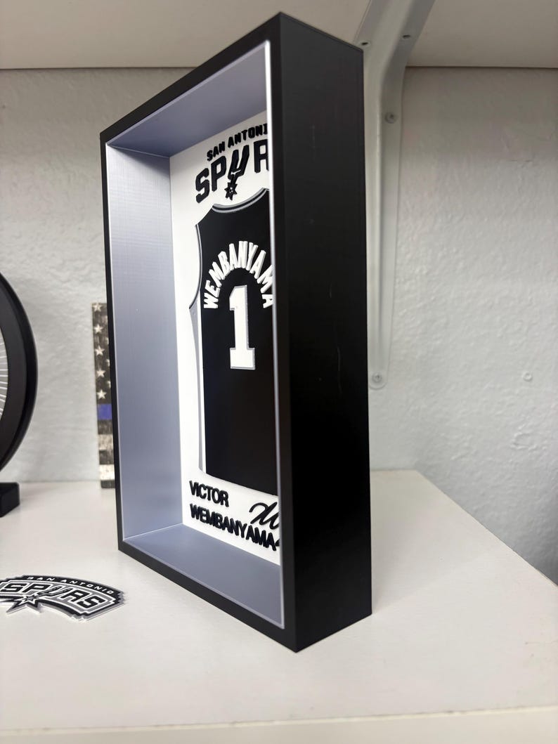 K&ouml;nnte beinhalten: Ein schwarzer Rahmen zeigt ein schwarz-wei&szlig;es San Antonio Spurs-Trikot mit dem Namen "Wembanyama" und der Nummer "1". Das Trikot wird in einer schwarzen Schattenbox mit wei&szlig;em und grauem Innenraum pr&auml;sentiert. Der Text "San Antonio Spurs" ist oben zu sehen.