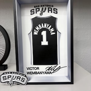 K&ouml;nnte beinhalten: Ein gerahmtes schwarz-wei&szlig;es San Antonio Spurs Trikot mit dem Namen "Wembanyama" und der Nummer "1". Der Rahmen enth&auml;lt das Teamlogo und die Unterschrift des Spielers. Ein Spurs-Logo-Aufkleber befindet sich im Vordergrund.