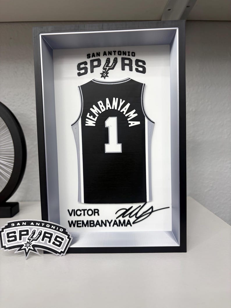 K&ouml;nnte beinhalten: Ein gerahmtes schwarz-wei&szlig;es San Antonio Spurs Trikot mit dem Namen "Wembanyama" und der Nummer "1". Der Rahmen enth&auml;lt das Teamlogo und die Unterschrift des Spielers. Ein Spurs-Logo-Aufkleber befindet sich im Vordergrund.
