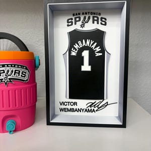 K&ouml;nnte beinhalten: Ein gerahmtes schwarz-wei&szlig;es San Antonio Spurs Trikot mit dem Namen "Wembanyama" und der Nummer "1". Der Rahmen enth&auml;lt den Teamnamen und eine Unterschrift. Links ist ein rosa-oranger Wasserk&uuml;hler zu sehen.