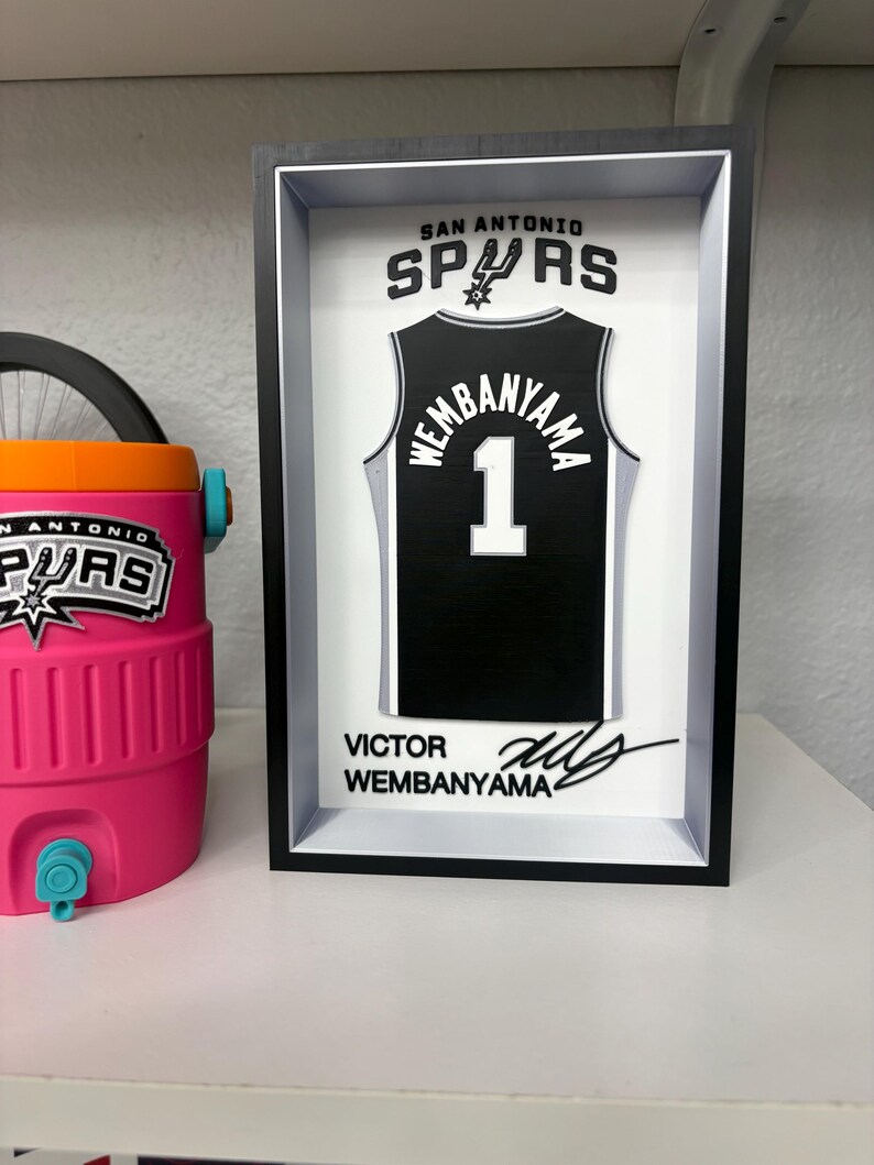 K&ouml;nnte beinhalten: Ein gerahmtes schwarz-wei&szlig;es San Antonio Spurs Trikot mit dem Namen "Wembanyama" und der Nummer "1". Der Rahmen enth&auml;lt den Teamnamen und eine Unterschrift. Links ist ein rosa-oranger Wasserk&uuml;hler zu sehen.