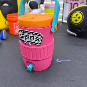 May include: A pink and orange San Antonio Spurs water cooler with a blue handle and spout. The cooler has the Spurs logo on the front and is likely a fan item.