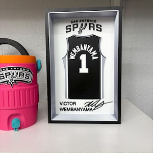 K&ouml;nnte beinhalten: Gerahmtes schwarz-wei&szlig;es San Antonio Spurs-Trikot mit "Wembanyama" und der Nummer "1". Der Rahmen enth&auml;lt das Teamlogo und die Unterschrift des Spielers. Links ist eine rosa-orangefarbene K&uuml;hlbox zu sehen.