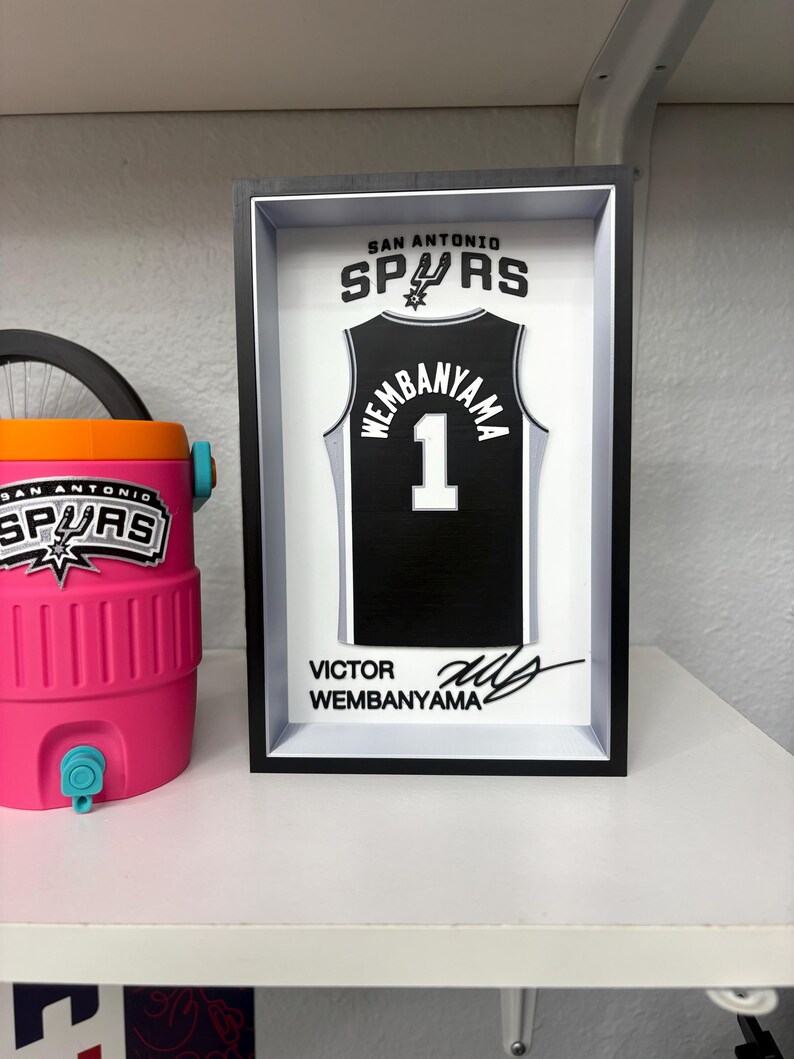 K&ouml;nnte beinhalten: Gerahmtes schwarz-wei&szlig;es San Antonio Spurs-Trikot mit "Wembanyama" und der Nummer "1". Der Rahmen enth&auml;lt das Teamlogo und die Unterschrift des Spielers. Links ist eine rosa-orangefarbene K&uuml;hlbox zu sehen.