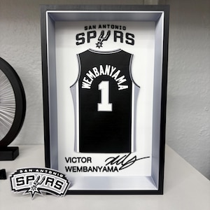 K&ouml;nnte beinhalten: Gerahmtes schwarz-wei&szlig;es San Antonio Spurs Trikot mit "Wembanyama" und der Nummer "1". Der Rahmen enth&auml;lt das Teamlogo und die Unterschrift des Spielers. Ein Spurs-Logo-Aufkleber ist ebenfalls vorhanden.