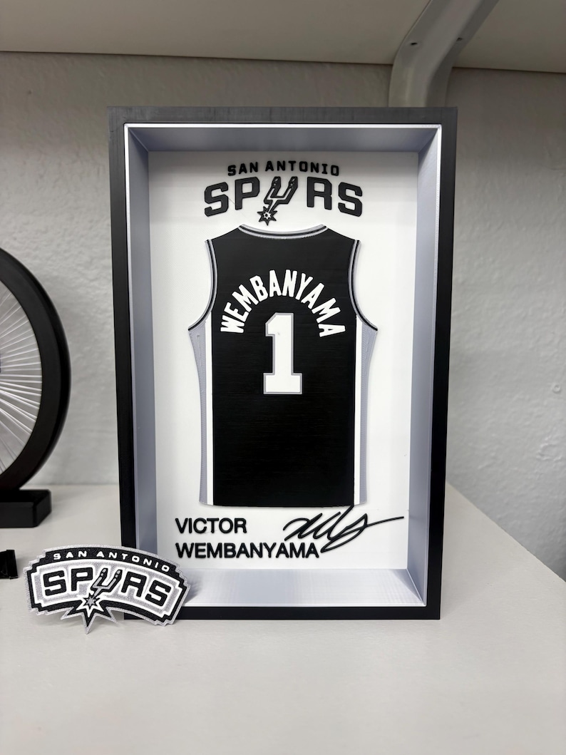 K&ouml;nnte beinhalten: Gerahmtes schwarz-wei&szlig;es San Antonio Spurs Trikot mit "Wembanyama" und der Nummer "1". Der Rahmen enth&auml;lt das Teamlogo und die Unterschrift des Spielers. Ein Spurs-Logo-Aufkleber ist ebenfalls vorhanden.