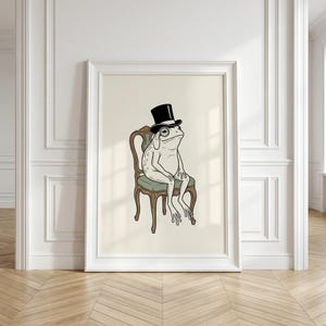 May include: A framed wall art print featuring a line-art illustration of a frog wearing a black top hat and a monocle, sitting on a wooden chair with a green upholstered seat. The frog is depicted in a seated position against a cream background.