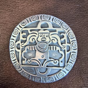 Op de afbeelding: Een ronde zilverkleurige metalen broche met een gestileerde centrale figuur met vloeiend haar en geometrische ledematen. De buitenrand is versierd met een herhalend patroon van concentrische cirkels en het geheel heeft een gepolijste afwerking.