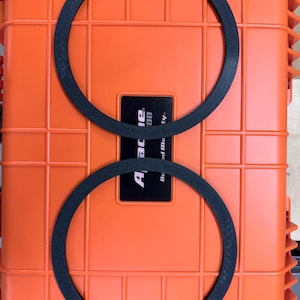 May include: Two black circular rings with a textured surface are positioned on top of a bright orange rectangular hard-shell case. The case features a black central label with the white text Apache and additional partially visible text.
