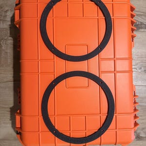 May include: A bright orange rectangular hard-shell protective case with a grid-like molded exterior texture. Two thin, black, circular foam gaskets with four small pre-drilled mounting holes each are positioned vertically on the top surface.