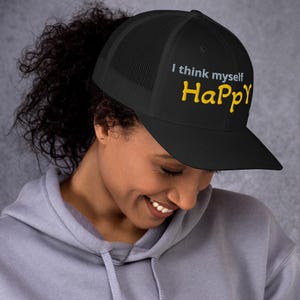 Könnte beinhalten: Schwarze Trucker-Mütze mit dem Text "I think myself Happy" in Weiß und Gelb. Die Mütze hat einen Mesh-Rücken und einen gebogenen Schirm. Die Mütze wird von einer Person getragen.
