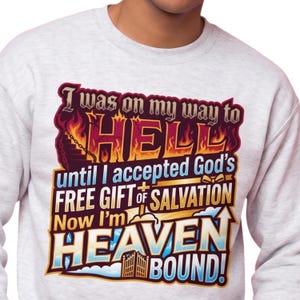 以下が含まれることがあります： ライトグレーのクルーネックトレーナー。前面には「I was on my way to HELL until I accepted God's FREE GIFT of SALVATION Now I'm HEAVEN BOUND!」というテキストと、炎、階段、十字架、門が描かれたグラフィックプリントが施されています。