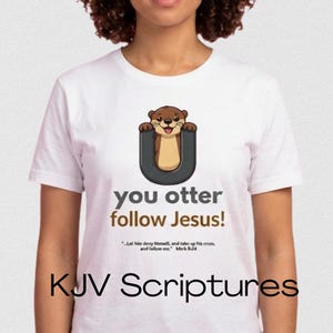 Op de afbeelding: Wit T-shirt met een cartoon otter in een donkergrijze "U" met de tekst "you otter follow Jesus!". Daaronder staat een bijbelvers en de woorden "KJV Scriptures".