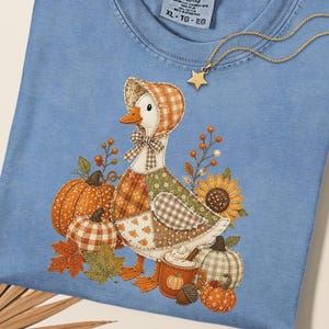 May include: A blue jean colored t-shirt featuring a graphic of a duck wearing a bonnet, surrounded by pumpkins, autumn leaves, a sunflower, and a mug of coffee. The shirt has a Comfort Colors label at the neck and a gold star pendant necklace.