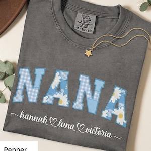 Puede incluir: Una camiseta gris Comfort Colors con la palabra "NANA" en letras azul claro con un estampado de cuadros y margaritas. Los nombres "hannah luna victoria" están debajo de la palabra "NANA". Un collar de estrella dorada está en la camiseta.