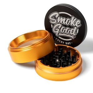 May include: A gold-colored, three-piece grinder with a black lid. The lid has the words "Smoke Good" in white script. The grinder is designed to break down herbs and spices. The grinder is made of metal.