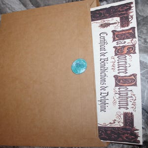May include: A brown paper envelope sealed with a turquoise wax seal. A decorative certificate with ornate lettering and illustrations is partially visible, with the text "Certificat de Bénédictions de Delphine".