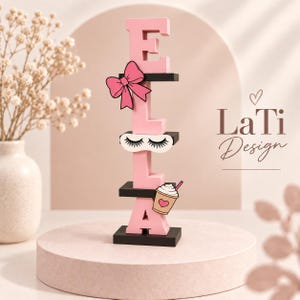 May include: A vertical wooden name sign spelling ELLA in pink block letters, separated by black horizontal shelves. The sign features a pink bow, a white sleep mask with black eyelashes, and a coffee cup graphic with a heart. Text reads LaTi Design.