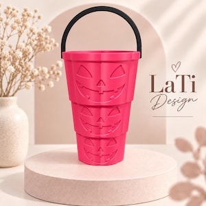 May include: A bright pink, tiered plastic bucket featuring three stacked jack-o'-lantern faces with carved eyes, noses, and wide grins. The bucket has a solid black arched handle attached to the top rim. The text LaTi Design is visible.