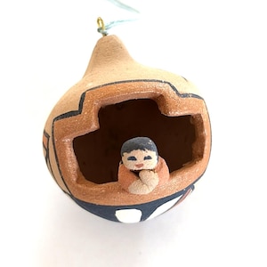 May include: A hollow, rounded terracotta ornament featuring a cutout opening with a small, hand-painted clay figure resting inside. The exterior is decorated with black and white geometric patterns and includes a small loop at the top for hanging.