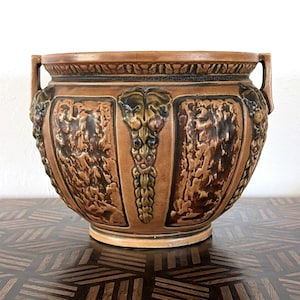 May include: A brown ceramic planter with a textured surface and decorative relief panels. The planter features handles and a detailed rim. The central panel has a grape vine design. The planter is a decorative item.