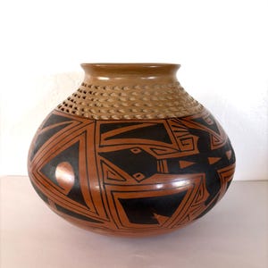 May include: A handcrafted, round pottery vase with a wide base and a narrow neck. The vase features a brown and black geometric design. The upper portion has a textured, light brown band with small holes. The vase is likely used for decorative purposes.