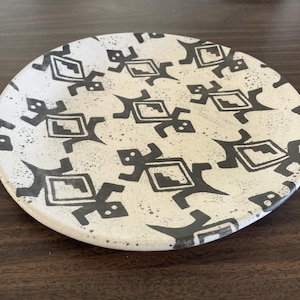May include: A round, off-white ceramic plate with a repeating black geometric design. The design features stylized figures with rectangular bodies and angular limbs. The plate has a speckled texture and a slightly raised edge.