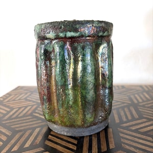 May include: A handmade ceramic tumbler with a textured, ribbed design. The cup features a blend of green, brown, and iridescent colors. The rim and base have a darker, textured appearance, adding to its unique, artistic appeal.