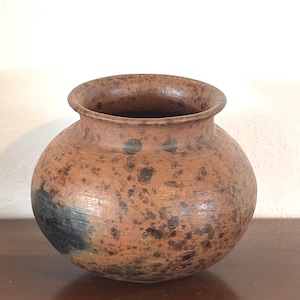 May include: A round, wide-mouthed earthenware pot with a textured surface. The vessel features a mottled brown and tan color palette with dark, irregular black spots and a prominent dark charcoal-colored patch on the lower left side of the body.