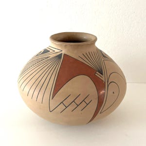 May include: A beige ceramic pot with a rounded body and a narrow opening. The pot is decorated with geometric designs in black and reddish-brown. The design includes lines, triangles, and curved shapes.