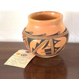 May include: A terracotta-colored ceramic jar featuring black geometric patterns and swirling line designs. A beige exhibition tag is attached, printed with "Museum of Northern Arizona," "Hopi Craftsman Exhibition," "July 2-6," "Anita Polacca," and "Jar."