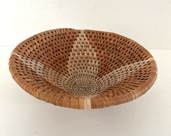 May include: A woven wicker bowl with a light brown and tan color pattern. The bowl is round and has a slight curve to the sides.