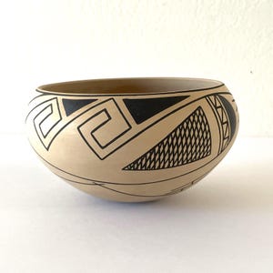 May include: A beige ceramic bowl with black geometric designs. The bowl features a series of black shapes, including triangles, squares, and curved lines. The interior of the bowl is a darker shade of beige. The bowl is round and has a wide opening.