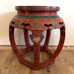 May include: A red, round, wooden stool with ornate floral and geometric designs. The stool features a red base with curved legs and a decorative top with a green border. The floral designs are in gold and white.