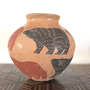 May include: A tan-colored ceramic vase with a rounded body and a wide opening. The vase features etched designs of bears in black and reddish-brown. The bears are depicted in various poses against a textured background.