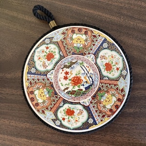 May include: A round ceramic plate with a black rope handle. The plate features a colorful Japanese-inspired design with flowers, geometric patterns, and gold accents.