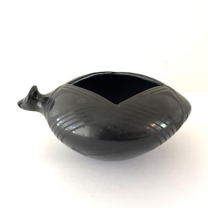 May include: A black ceramic bowl with a unique, asymmetrical shape. The bowl features a square opening and a small, pointed extension on one side. Diagonal lines are etched into the surface, adding texture. The bowl is likely handmade and suitable for decorative use.