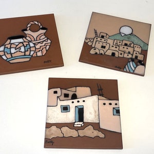 May include: Three ceramic tiles with Southwestern-inspired designs. The first tile features two brown and blue pottery vessels. The second tile depicts a village of adobe houses with a green hill and a white sun in the background. The third tile shows a single adobe house with a ladder and a brown door. The tiles are signed "Betty" and "Bette".