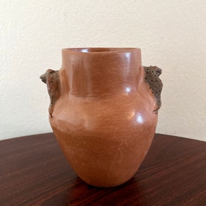 May include: A terracotta pottery vase with two sculpted frog handles. The vase is a reddish-brown color and has a smooth, slightly textured surface.