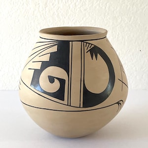 May include: A beige ceramic pot with a rounded body and a wide opening. The pot is adorned with a bold black geometric design featuring curved and linear elements. The rim is a darker shade of brown. This handcrafted piece showcases traditional pottery.
